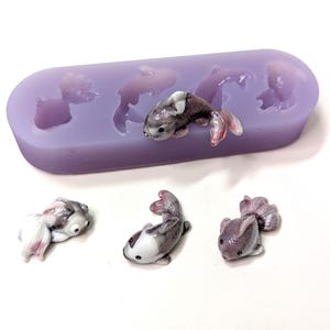 K&ouml;nnte beinhalten: Eine lavendelfarbene Silikonform mit fischf&ouml;rmigen Hohlr&auml;umen, zusammen mit mehreren fertigen Miniatur-Fischfiguren. Die Figuren sind wei&szlig;, grau und rosa, mit detaillierten Flossen und Schuppen. Die Form ist rechteckig mit abgerundeten Kanten.