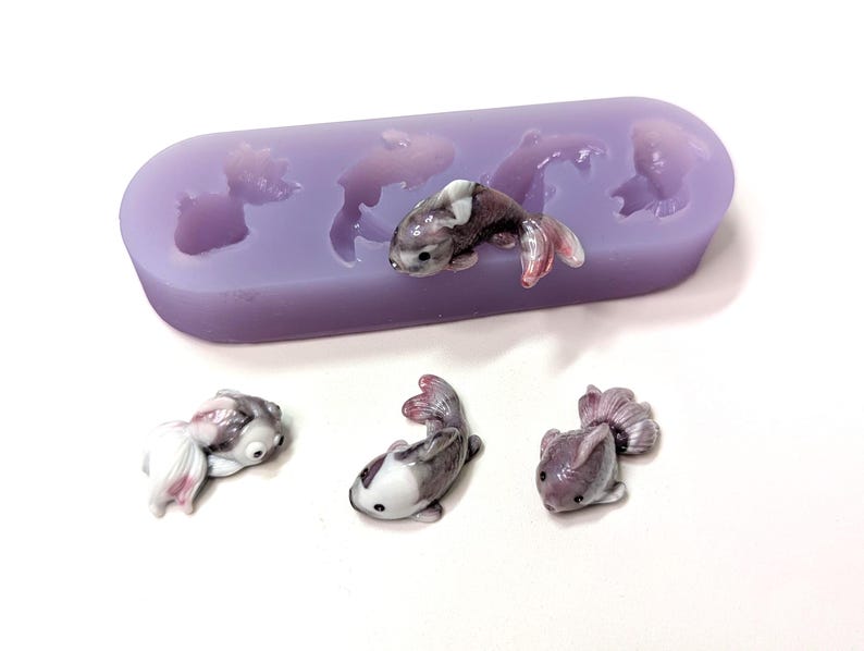 K&ouml;nnte beinhalten: Eine lavendelfarbene Silikonform mit fischf&ouml;rmigen Hohlr&auml;umen, zusammen mit mehreren fertigen Miniatur-Fischfiguren. Die Figuren sind wei&szlig;, grau und rosa, mit detaillierten Flossen und Schuppen. Die Form ist rechteckig mit abgerundeten Kanten.