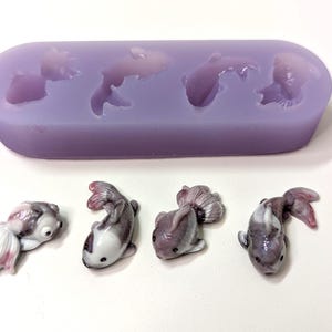 K&ouml;nnte beinhalten: Eine lavendelfarbene Silikonform mit vier Goldfisch-Abdr&uuml;cken, neben vier fertigen Miniatur-Goldfischfiguren. Die Figuren sind wei&szlig;, grau und rosa, mit detaillierten Flossen und Augen. Die Form ist rechteckig und die Figuren liegen auf einer wei&szlig;en Oberfl&auml;che.