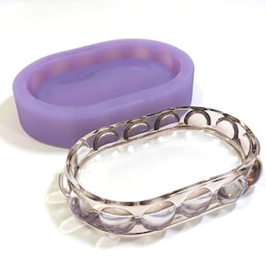 Puede incluir: Un molde ovalado de silicona de color lavanda junto a una bandeja decorativa ovalada de color plateado. La bandeja tiene una base de vidrio transparente y un borde decorativo con recortes semicirculares. Los artículos están sobre una superficie blanca.