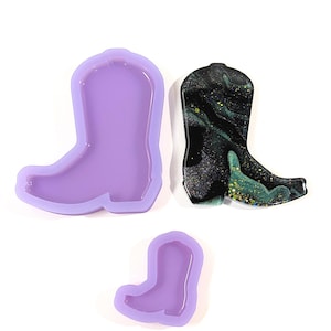 May include: Three items: a purple silicone mould in the shape of a cowboy boot, a finished boot with a black, green, and gold marbled design, and a smaller purple silicone mould of a boot. All items are on a white background.