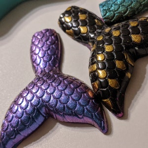 May include: Two mermaid tail charms, one is iridescent purple and the other is black with gold accents.
