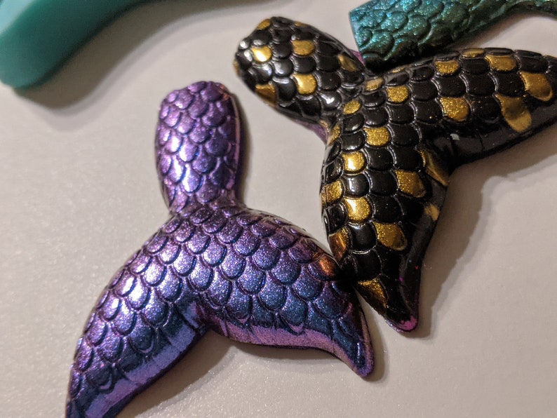 May include: Two mermaid tail charms, one is iridescent purple and the other is black with gold accents.
