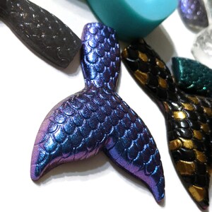 May include: A purple and blue iridescent mermaid tail charm with a scaly texture. The charm is made of resin and is perfect for adding a touch of whimsy to jewelry or crafts.