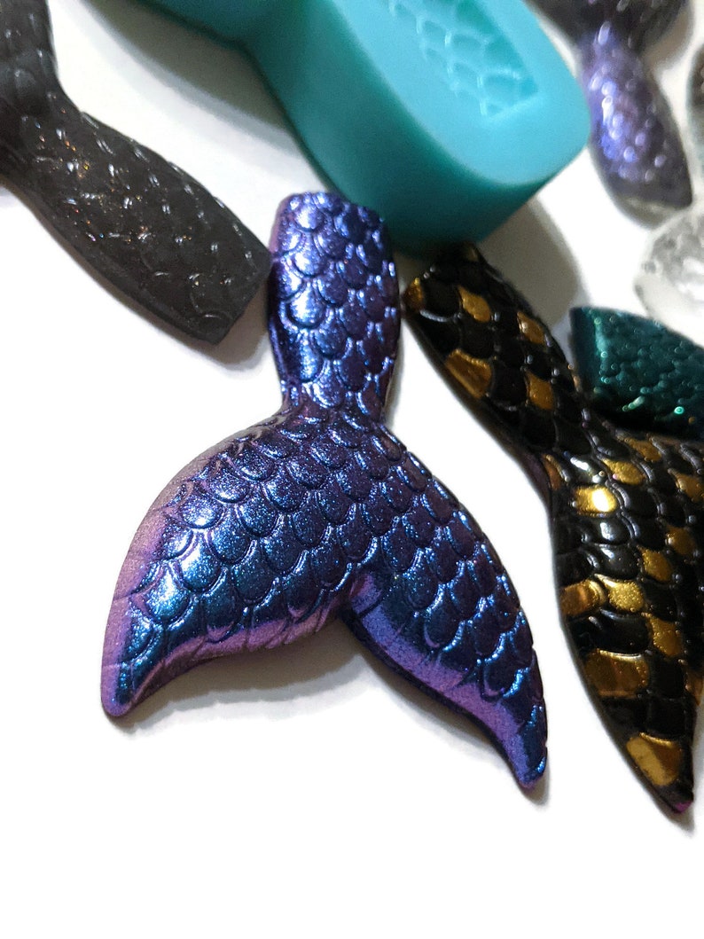May include: A purple and blue iridescent mermaid tail charm with a scaly texture. The charm is made of resin and is perfect for adding a touch of whimsy to jewelry or crafts.