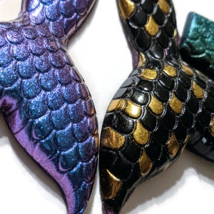 May include: Two mermaid tail charms, one with iridescent purple and blue scales and the other with black and gold scales.