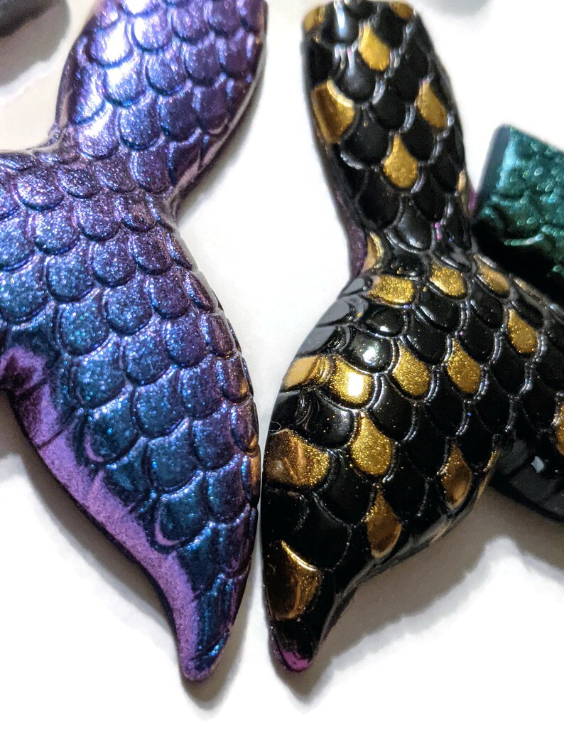 May include: Two mermaid tail charms, one with iridescent purple and blue scales and the other with black and gold scales.