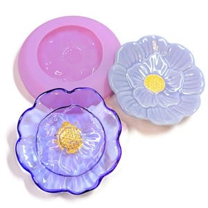 May include: A collection of flower-shaped items. A lavender silicone mold sits next to two finished flower-shaped objects. One is a light blue ceramic flower with a yellow center, and the other is a transparent purple glass flower with a gold center.