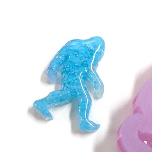 K&ouml;nnte beinhalten: Hellblaue, glitzernde Kunstharzfigur eines Bigfoot beim Gehen. Die Figur hat eine durchscheinende Qualit&auml;t und befindet sich vor einem wei&szlig;en Hintergrund. Ein lavendelfarbenes Objekt ist teilweise sichtbar.