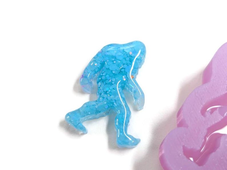K&ouml;nnte beinhalten: Hellblaue, glitzernde Kunstharzfigur eines Bigfoot beim Gehen. Die Figur hat eine durchscheinende Qualit&auml;t und befindet sich vor einem wei&szlig;en Hintergrund. Ein lavendelfarbenes Objekt ist teilweise sichtbar.