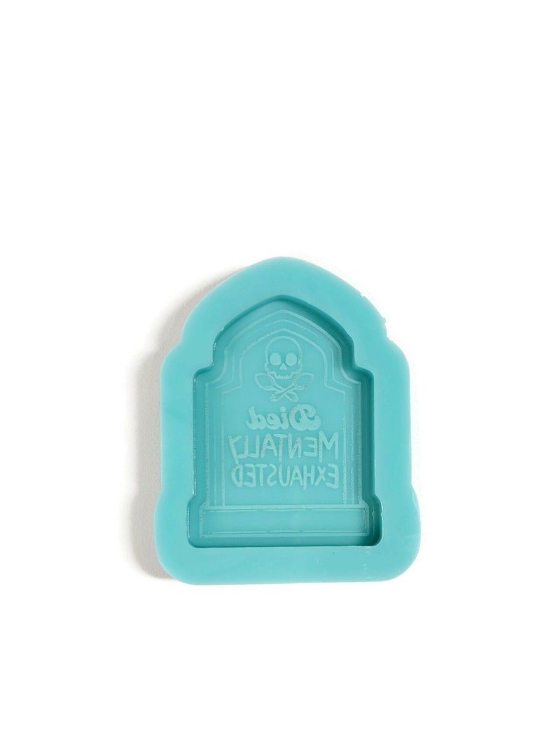 Puede incluir: Un molde de silicona azul claro con forma de l&aacute;pida con una calavera y huesos cruzados y el texto "EXHAUSTED MENTALLY Died".