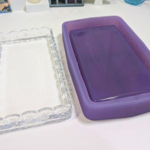 May include: A clear, rectangular glass tray with a scalloped edge and floral detailing sits next to a purple silicone mold filled with a glossy, lavender-colored substance. The tray is empty, and the mold is full.