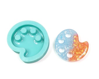 Könnte beinhalten: Eine tealfarbene Silikonform in Form einer Farbpalette mit fünf kreisförmigen Vertiefungen. Neben der Form befindet sich eine fertige Harz-Farbpalette mit einem blauen und orangefarbenen Swirl-Design.