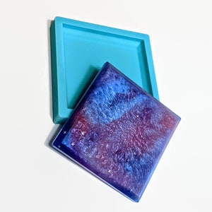May include: A square resin coaster with a galaxy design in shades of blue, purple, and white, with small star accents. A turquoise silicone mould is next to the coaster. The coaster is a handmade item.