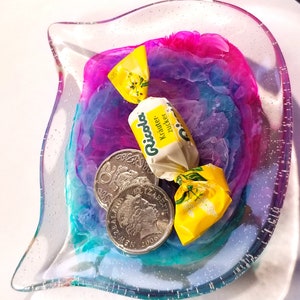 May include: A clear glass dish with a pink, blue, and purple swirl design. The dish contains two silver coins and a yellow wrapped candy with the text "Ricola Kr&auml;uter Zucker" on it.
