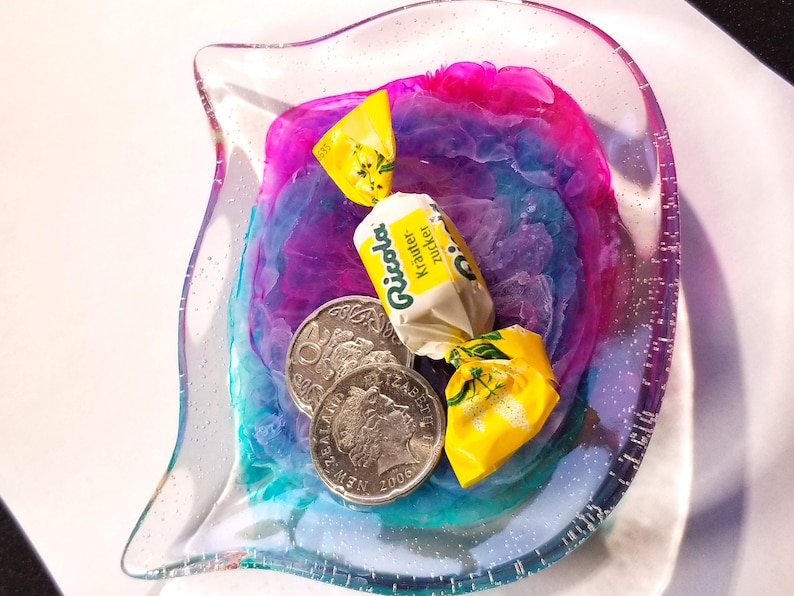 May include: A clear glass dish with a pink, blue, and purple swirl design. The dish contains two silver coins and a yellow wrapped candy with the text "Ricola Kr&auml;uter Zucker" on it.