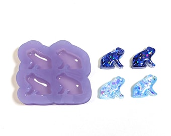 May include: A purple silicone mold with four frog-shaped cavities, next to four finished frog resin charms. The charms are blue with glitter, in various shades. The frogs are in a sitting position.