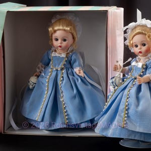Könnte beinhalten: Zwei Vintage-Porzellanpuppen in einer Box. Die Puppen tragen hellblaue Kleider mit goldenen Verzierungen und weiße Spitzenhauben. Eine Puppe befindet sich in der Box, die andere steht draußen und hält eine kleine Handtasche. Die Box ist hellblau mit rosa Punkten.