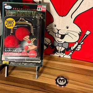 May include: A red, white, and black magic trick set with a package that says "World Greatest Magic" and "2005 Latest Work". The package shows a magician holding three red sponge balls. The package is on a wooden stand. A red and white rabbit with a black bow tie and a black wand is on a red background. A black and pink poker chip with the word "Magic" on it is on the wooden surface.