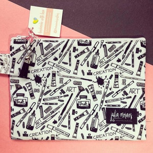 May include: A white zippered pouch with a black and white print of art supplies, including paintbrushes, paint tubes, and the words "Art" and "Creation". The pouch also has a black label with the text "julia Moraes".
