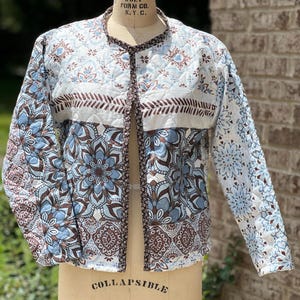 May include: A quilted jacket with a floral pattern in shades of blue, brown, and white. The jacket is displayed on a dress form. The collar and trim are a dark brown with a small floral design. The jacket has long sleeves.