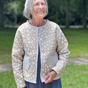 May include: A beige quilted jacket with a floral pattern and two front pockets. The jacket has a white and blue floral design and a contrasting patterned trim. The jacket is open, revealing a blue dress underneath.