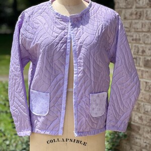May include: A light purple quilted jacket with a round collar and two front pockets. The jacket has a quilted pattern and is displayed on a dress form. The pockets have a floral pattern. The jacket is open in the front.