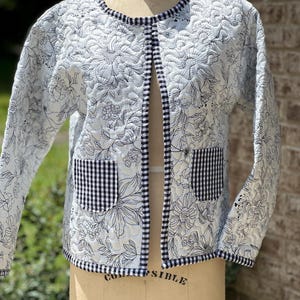May include: A white quilted jacket with a floral pattern in black. The jacket has a black and white gingham trim and two front pockets. The jacket is displayed on a mannequin.