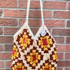 May include: A handmade crochet tote bag featuring a granny square design. The bag showcases a vibrant color palette of brown, yellow, and red, with cream-colored accents. It has two cream-colored straps and a small tag attached.