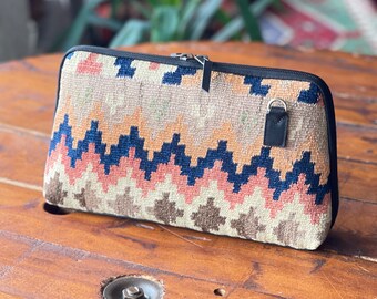 May include: A beige, pink, and blue patterned zippered pouch with a leather strap. The pouch is made of a woven fabric and has a geometric design.