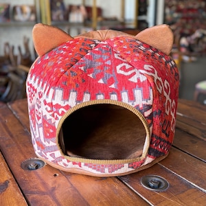 May include: A red, brown and white patterned cat bed with a round opening and two cat ears on top. The bed is made of fabric and has a soft, plush lining.