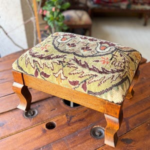 May include: A small, rectangular wooden footstool with a patterned fabric top in shades of green, brown, and orange. The footstool has four curved legs and is placed on a wooden surface.