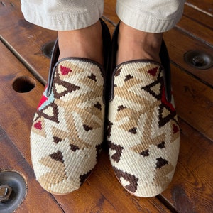 K&ouml;nnte beinhalten: Ein Paar beige und braune gewebte Slipper mit geometrischem Muster. Die Schuhe sind aus einem gewebten Stoff gefertigt und haben einen braunen Lederbesatz.