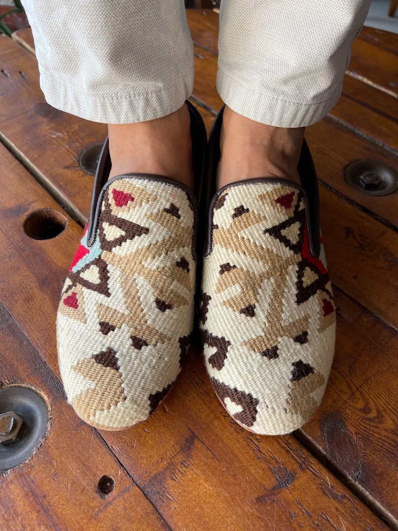 K&ouml;nnte beinhalten: Ein Paar beige und braune gewebte Slipper mit geometrischem Muster. Die Schuhe sind aus einem gewebten Stoff gefertigt und haben einen braunen Lederbesatz.
