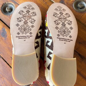 K&ouml;nnte beinhalten: Ein Paar braune und wei&szlig;e gemusterte Hausschuhe mit Ledersohlen. Die Sohlen haben ein geometrisches Muster und der Text "Vintage handwoven kilim upper Genuine leather sole 41" ist auf der Unterseite aufgedruckt.