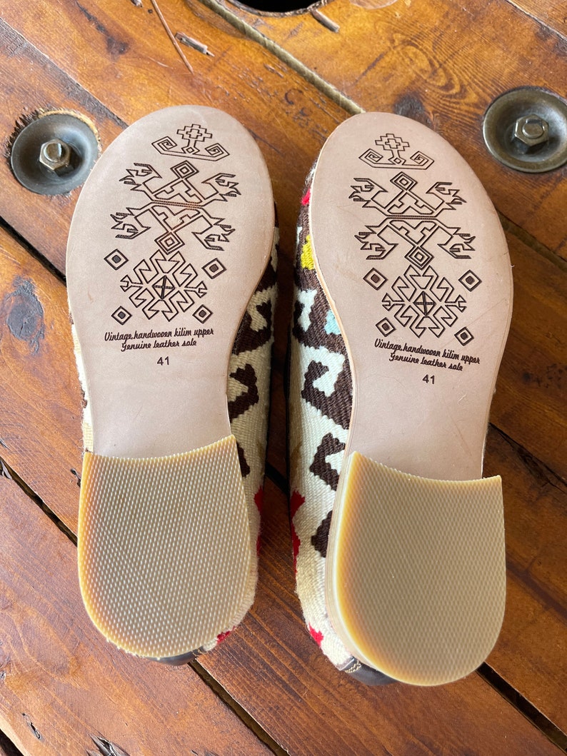 K&ouml;nnte beinhalten: Ein Paar braune und wei&szlig;e gemusterte Hausschuhe mit Ledersohlen. Die Sohlen haben ein geometrisches Muster und der Text "Vintage handwoven kilim upper Genuine leather sole 41" ist auf der Unterseite aufgedruckt.