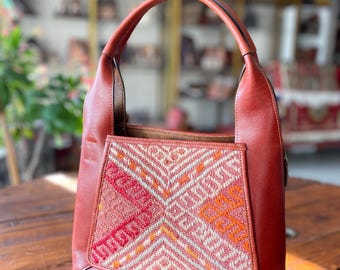 Op de afbeelding: Een roodbruine leren tote bag met een geweven, geometrisch patroon op de voorzak. De tas heeft een enkele handgreep en staat op een houten oppervlak. Het geweven patroon heeft tinten rood, oranje en crème.