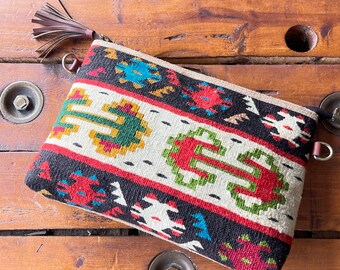May include: A colorful woven clutch bag with a geometric pattern in shades of red, green, blue, and yellow. The bag has a brown leather zipper pull and a tassel.