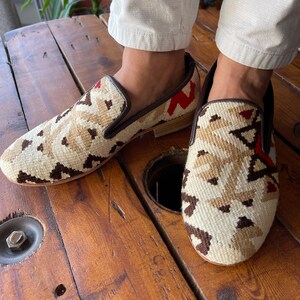 K&ouml;nnte beinhalten: Ein Paar braune Lederschuhe mit einem bunten geometrischen Muster. Die Schuhe sind aus einem gewebten Stoff mit roten, wei&szlig;en und braunen Akzenten. Die Schuhe werden von einer Person getragen, die eine wei&szlig;e Hose tr&auml;gt.