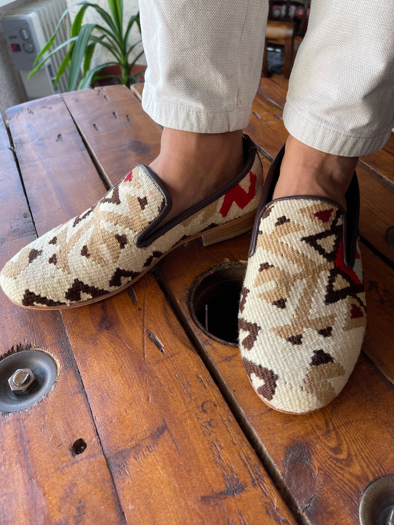 K&ouml;nnte beinhalten: Ein Paar braune Lederschuhe mit einem bunten geometrischen Muster. Die Schuhe sind aus einem gewebten Stoff mit roten, wei&szlig;en und braunen Akzenten. Die Schuhe werden von einer Person getragen, die eine wei&szlig;e Hose tr&auml;gt.