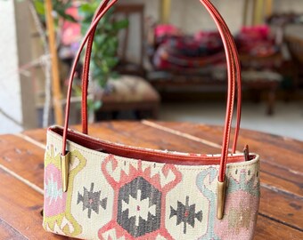 Op de afbeelding: Een rechthoekige handtas met een geweven textielontwerp in crème, roze, zwart en blauw. De tas heeft rode handvatten en goudkleurige metalen accenten. De tas staat op een houten tafel.
