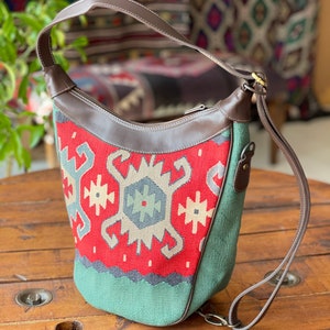 Op de afbeelding: Een rode, groene en witte gepatroneerde tote bag met een bruine leren band en handvat. De tas heeft een ritssluiting en een decoratieve metalen gesp.