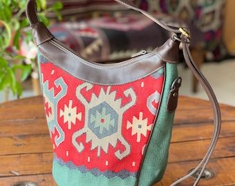 Op de afbeelding: Een rode, groene en witte gepatroneerde tote bag met een bruine leren band en handvat. De tas heeft een ritssluiting en een decoratieve metalen gesp.