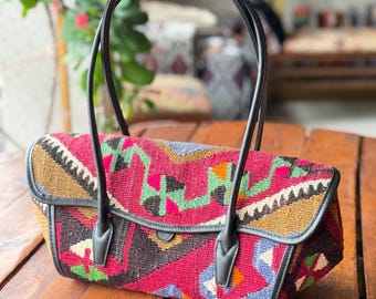 May include: A black leather tote bag with a colorful woven fabric design. The bag has two black leather handles and a flap closure.