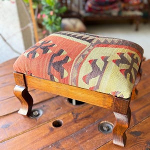 May include: A small, wooden footstool with a rectangular top and four curved legs. The top is upholstered with a patterned fabric in shades of orange, brown, and cream. The wooden frame has a warm, brown finish.