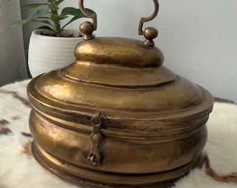 Puede incluir: Una caja vintage de latón con forma redondeada y tapa con bisagras. La caja tiene un asa decorativa en la parte superior y un cierre en la parte delantera.