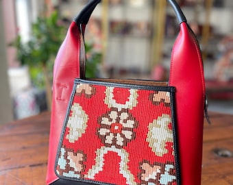 May include: A red leather tote bag with a black leather trim and a colorful patterned fabric insert. The bag has a single top handle.