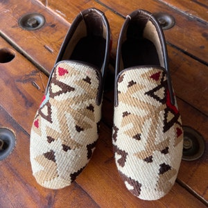 K&ouml;nnte beinhalten: Ein Paar braune Slipper aus Leder mit einem wei&szlig;en und braunen geometrischen Muster.
