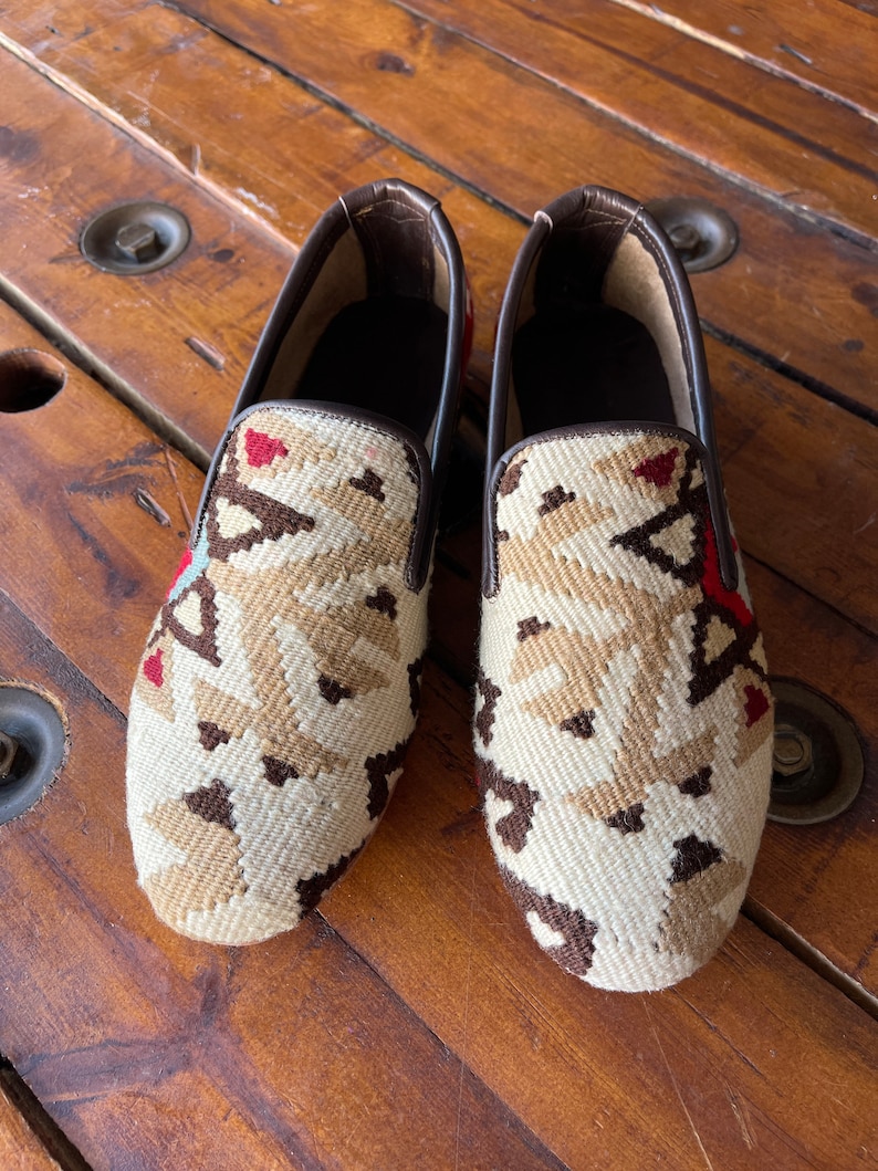 K&ouml;nnte beinhalten: Ein Paar braune Slipper aus Leder mit einem wei&szlig;en und braunen geometrischen Muster.