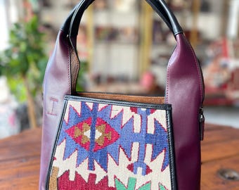 May include: A burgundy leather tote bag with a black leather handle and a zipper closure. The front of the bag features a colorful geometric pattern.